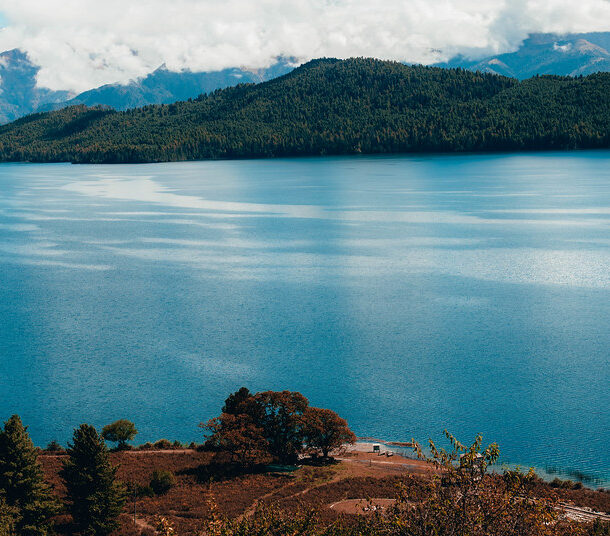 Kathmandu to Rara Lake Jeep Tour through serene blue lake and forested Himalayan hills under cloudy peaks.