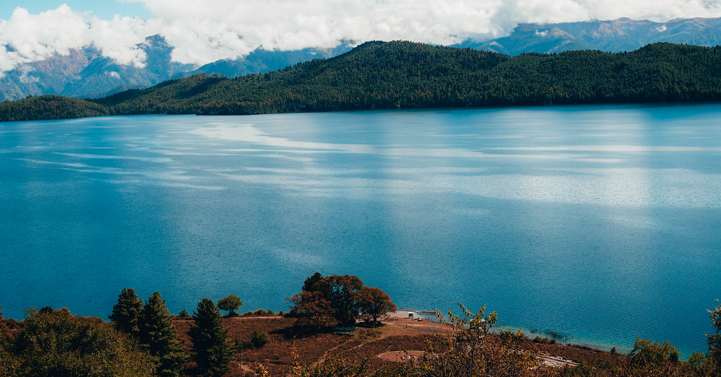 Kathmandu to Rara Lake Jeep Tour through serene blue lake and forested Himalayan hills under cloudy peaks.